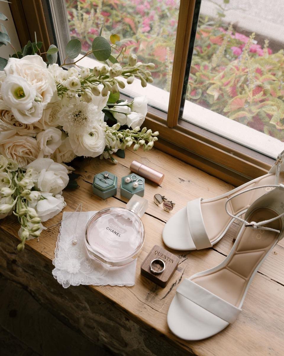 White wedding shoes and rings with a floral bouquet on a wooden windowsill.