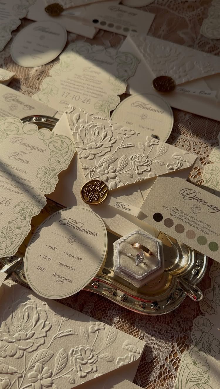 Wedding rings in a box surrounded by cream embossed invitations and lace fabric.