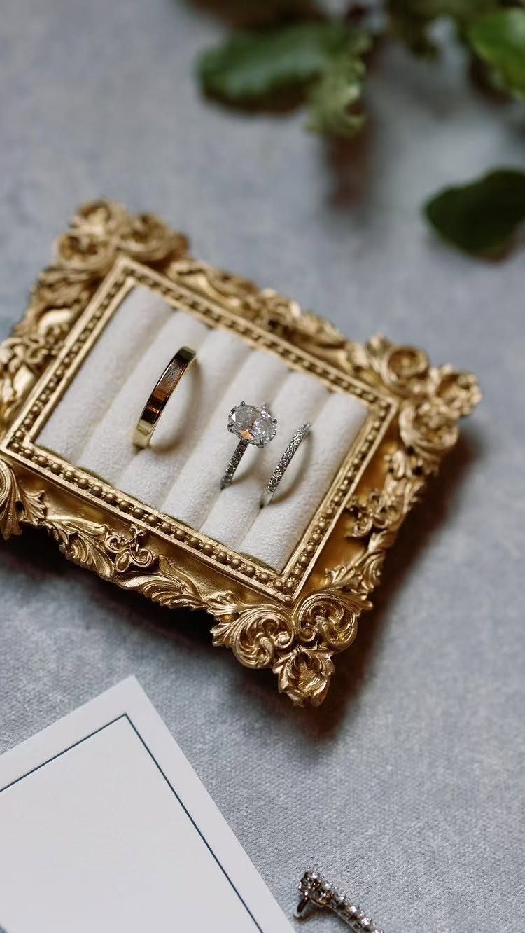 Three wedding rings in an ornate gold frame with a white card and leaves.