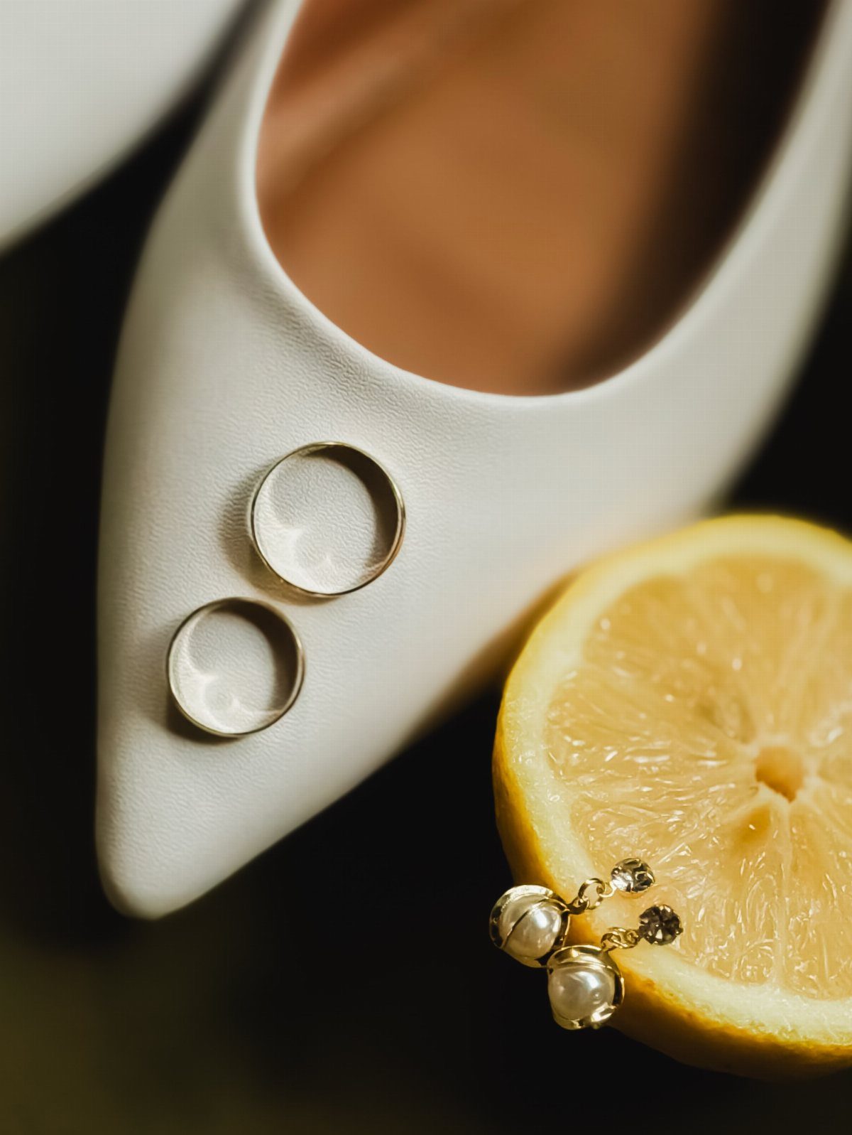 Two gold wedding rings resting on a white pointed-toe shoe beside a lemon slice.