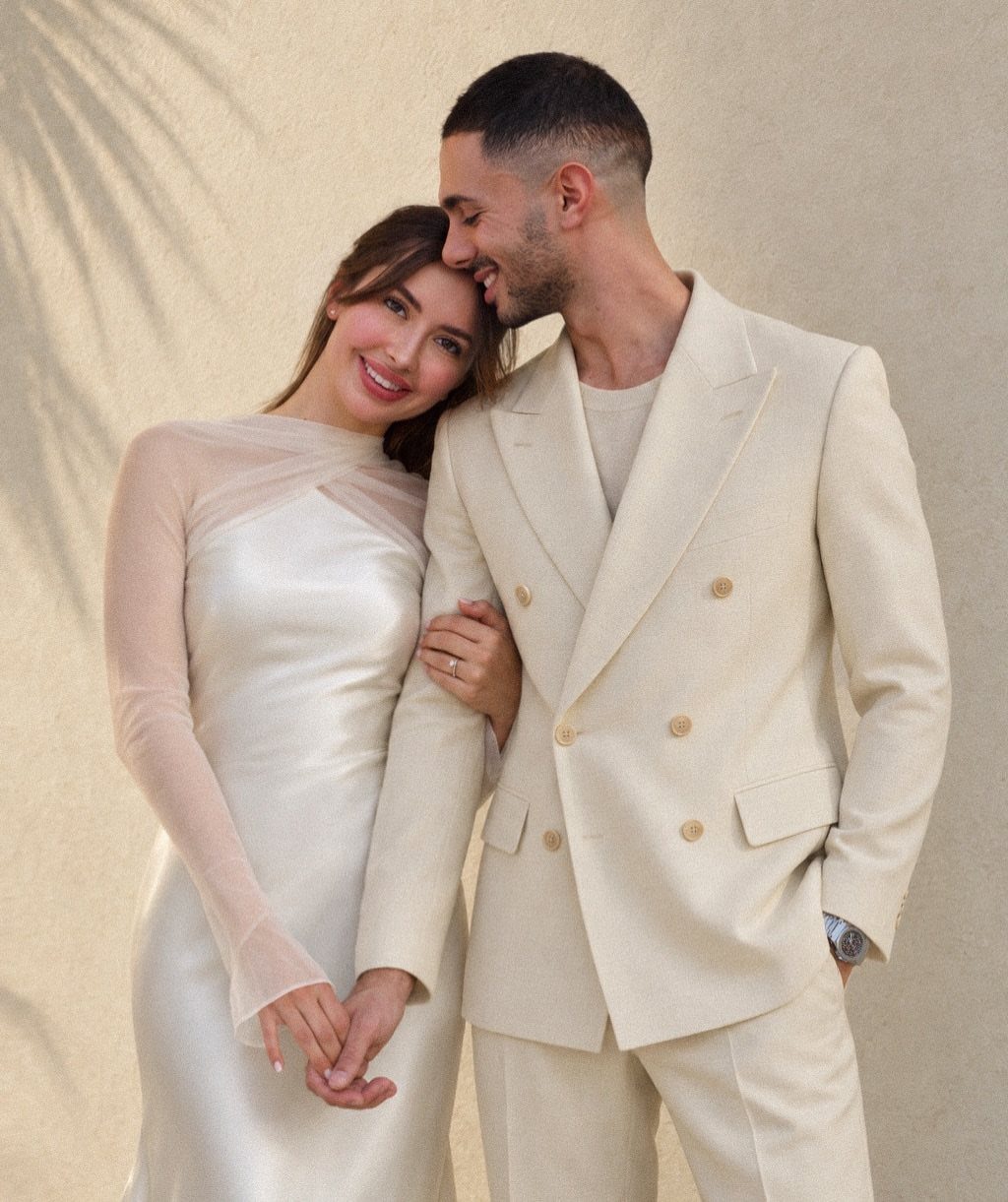 Couple in elegant white and cream formal attire posing against a beige wall