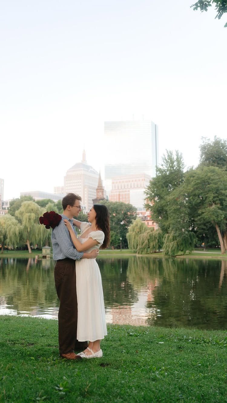 Couple embracing by a pond with a city skyline in the background.