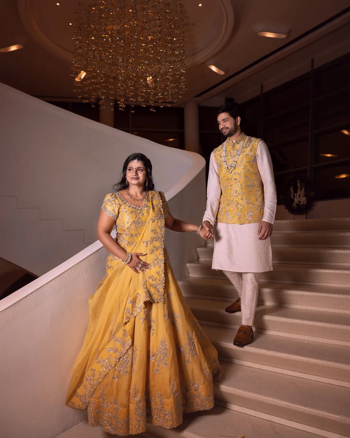 Couple in yellow and white traditional Indian attire posing on a white staircase.