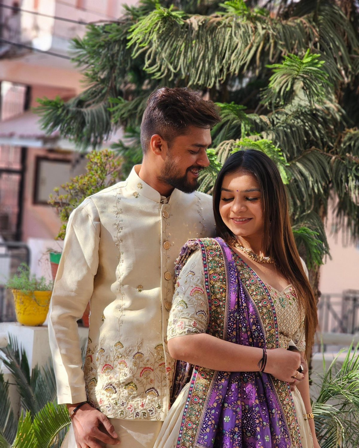 Couple in traditional Indian cream and purple attire posing outdoors near green trees.
