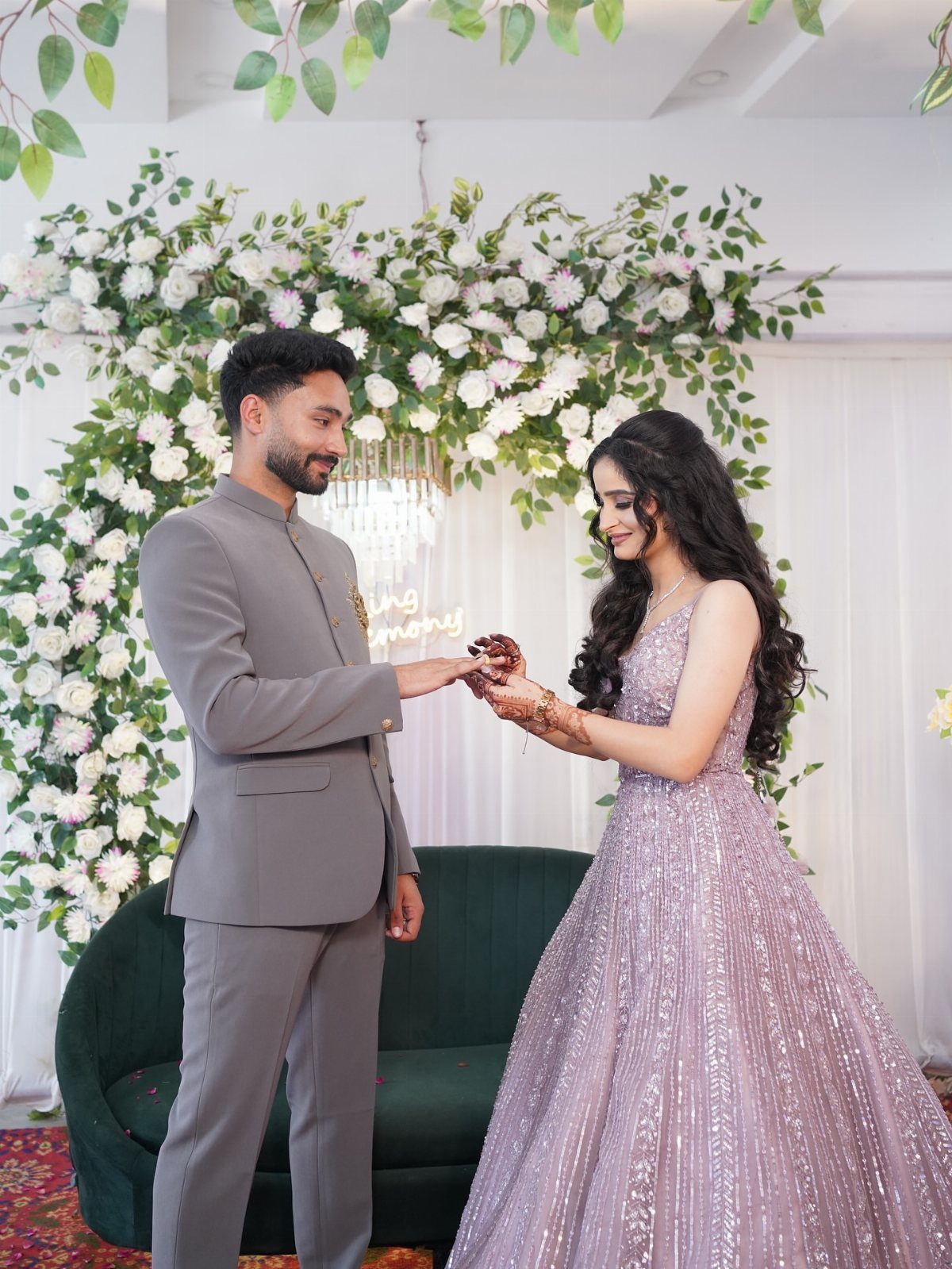 Couple in formal grey suit and lavender gown posing before a floral backdrop.