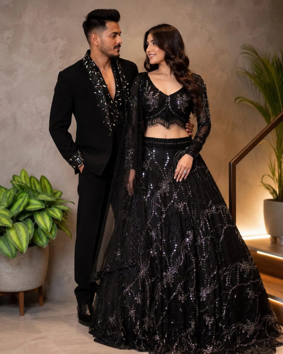 Couple posing in formal black sequined Indian attire against a grey wall.