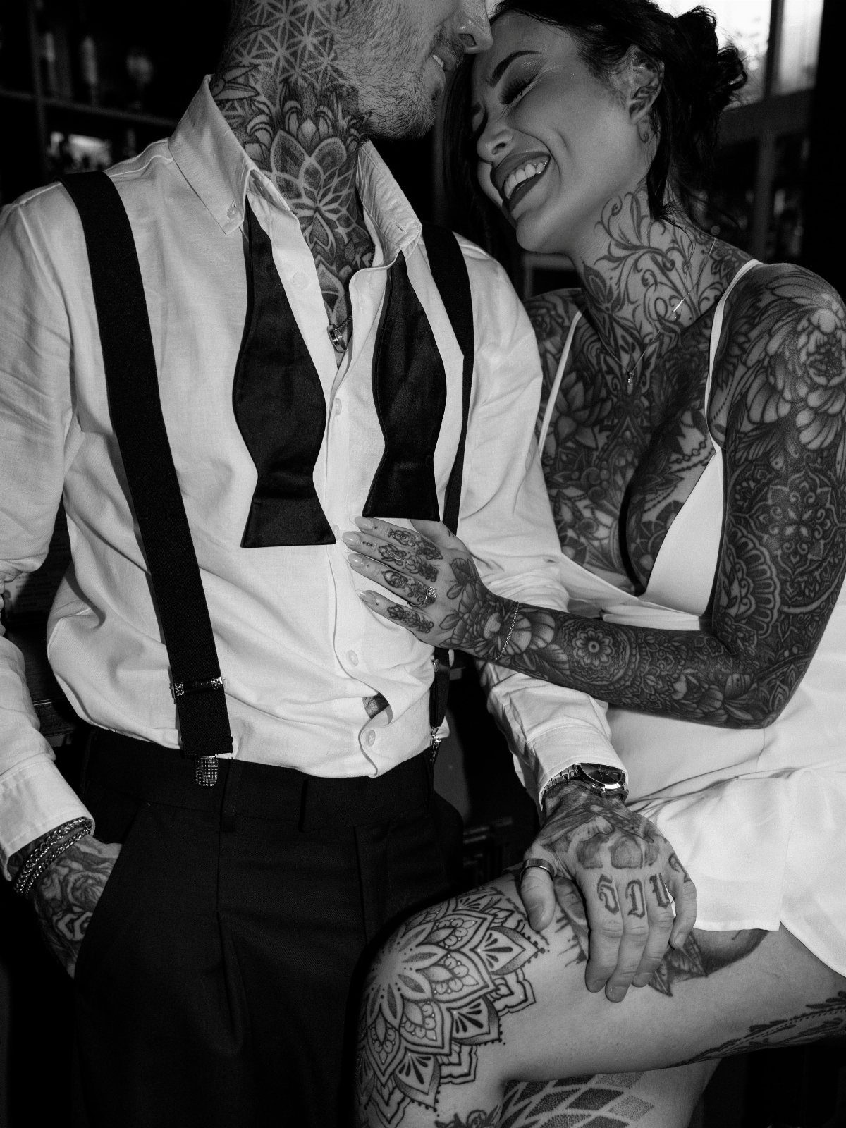 Tattooed couple in white formal attire smiling and posing together in black and white.