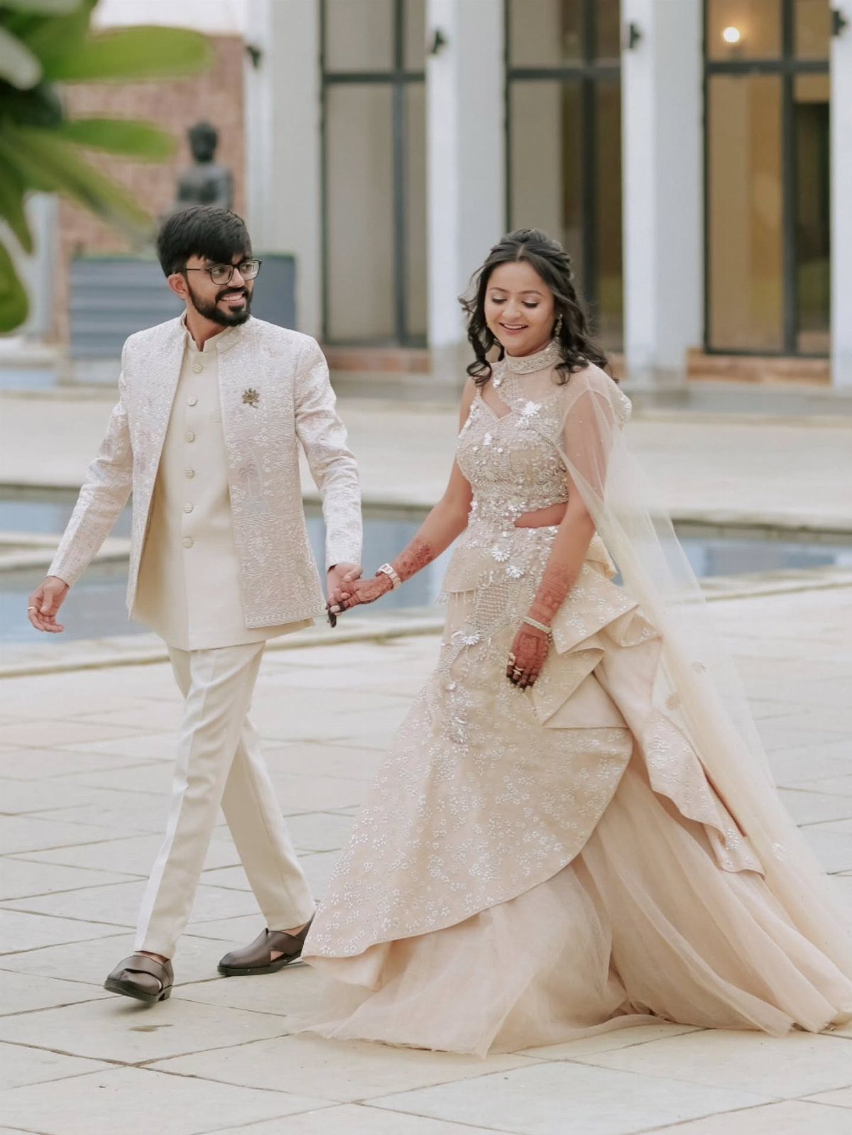 Couple in ivory and cream formal attire walking hand-in-hand on a tiled patio.