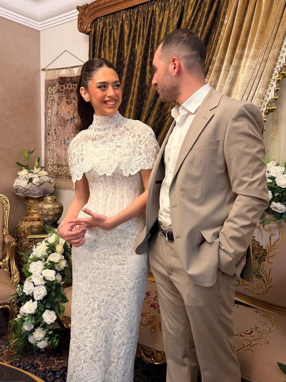 Woman in a white intricate lace dress and man in a tan suit posing.