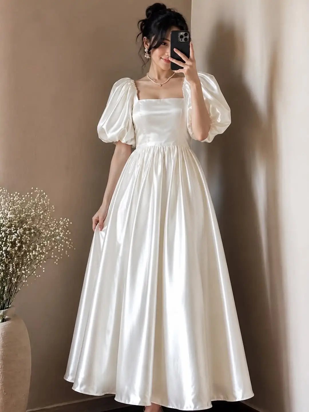 Woman in a long white satin puff sleeve dress taking a mirror selfie.